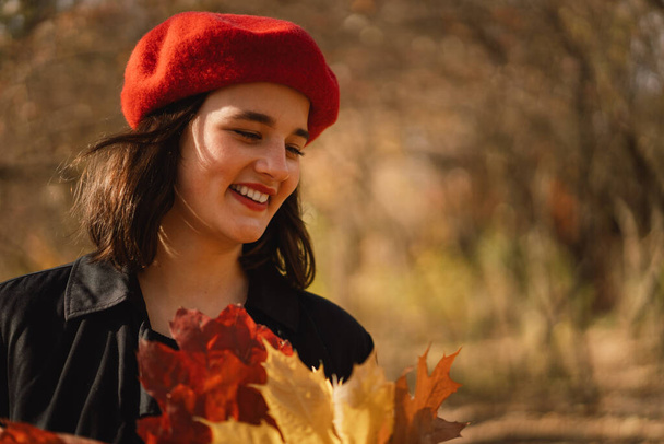 Una adolescente en una boina roja con un ramo de hojas de otoño en sus manos camina por el bosque - Foto, Imagen
