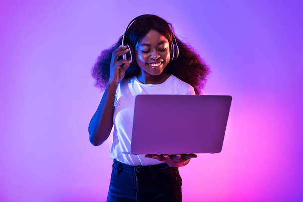 Νεαρή μαύρη γυναίκα που έχει βιντεοκλήση, χρησιμοποιεί φορητό υπολογιστή, φοράει ασύρματα ακουστικά σε νέον φως - Φωτογραφία, εικόνα