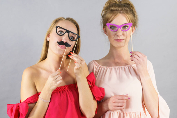 Happy two women holding paper decoration photo booth mask glasses and moustache on stick, having fun. Wedding, birthday and carnival funny accessories concept. - Фото, изображение
