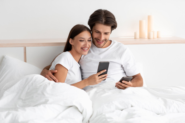 Iloinen valkoihoinen aviomies ja vaimo katselee älypuhelimia, halaa, katso video yhdessä sängyssä - Valokuva, kuva