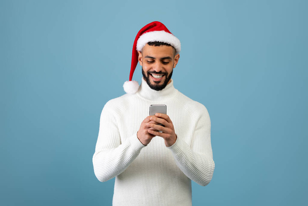 Wow. Glücklicher arabischer Typ mit Handy, Weihnachtsmütze und Lächeln auf blauem Studiohintergrund - Foto, Bild