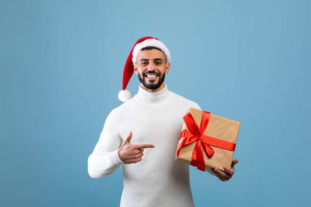 Geschenkübergabe. Glücklicher arabischer Mann mit Weihnachtsmütze, umwickelte Geschenkschachtel und Zeigefinger darauf, blauer Hintergrund - Foto, Bild