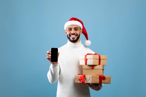 Mockup for Christmas ad. Happy arab man showing smartphone with blank screen and holding gifts, blue background - Fotoğraf, Görsel