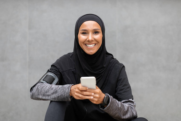 Szczęśliwa młoda muzułmanka w odzieży sportowej relaks ze smartfonem po treningu na świeżym powietrzu - Zdjęcie, obraz