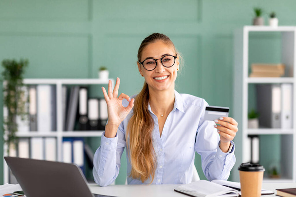 Könyvelő bankár hölgy kezében hitelkártya és mutató rendben jel, miközben ül a munkahelyen, mosolygós kamera az irodában - Fotó, kép