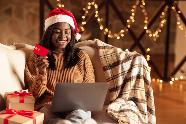 Online kerstinkopen doen. Zwarte vrouw in Santa hoed zitten in de buurt van geschenkdozen, met creditcard en met behulp van laptop thuis - Foto, afbeelding