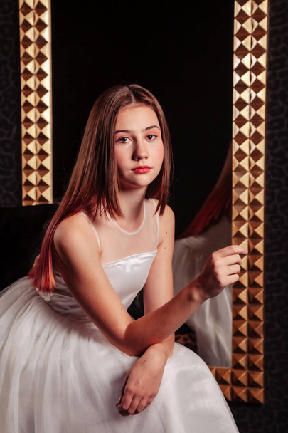 Portrait of pretty cute teenager girl in an elegant dress at mirror in dark stylish interior of living room. Children emotions and posing. Concept of style, fashion and beauty. Copy space for site - 写真・画像