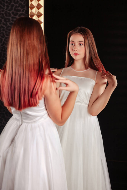 Portrait of pretty cute teenager girl in an elegant dress at mirror in dark stylish interior of living room. Children emotions and posing. Concept of style, fashion and beauty. Copy space for site - Photo, image