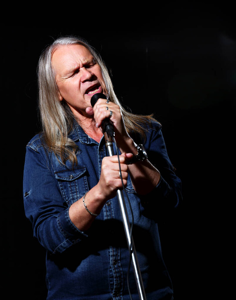 elderly man with long gray hair in blue denim shirt sings into a microphone on stage on black background - Photo, Image