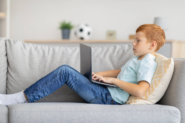 Punapää poika käyttää kannettavaa tietokonetta, pelaa online videopeli, sivunäkymä - Valokuva, kuva
