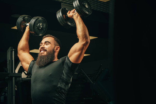 Bodybuilder doing exercises with dumbbells in a dark gym - Foto, Imagen