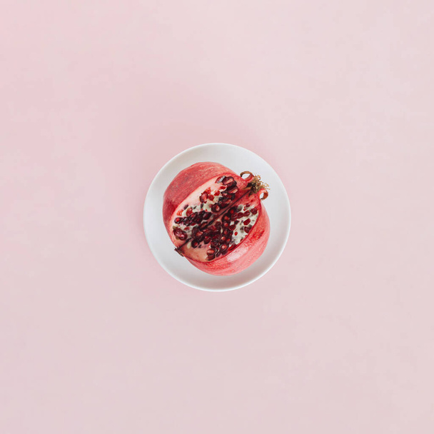 Summer fruit layout made of bold red cut pomegranate standing in the white plate isolated on the pastel pink background. Minimal Mediterranean flat lay. Raw organic vegetarian healthy food concept. - Foto, afbeelding