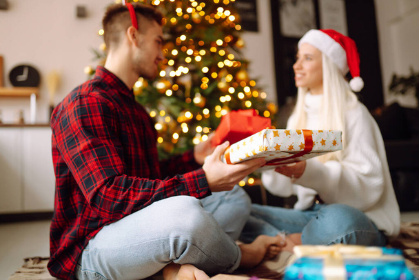 Exchange of Christmas gifts. Young couple opening gift box in cozy living room near Christmas tree. Romantic day. Winter holidays. - Foto, Imagem
