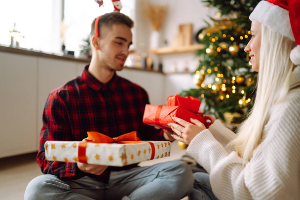 Exchange of Christmas gifts. Young couple opening gift box in cozy living room near Christmas tree. Romantic day. Winter holidays. - 写真・画像
