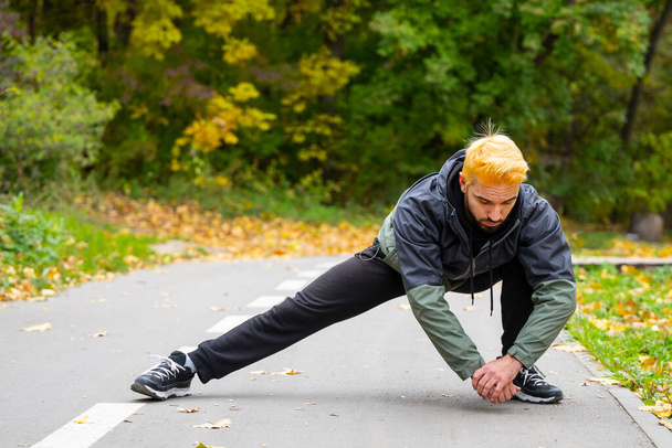 Man with dyed hair in blonde doing stretching at the city park. Open air training. - Фото, зображення