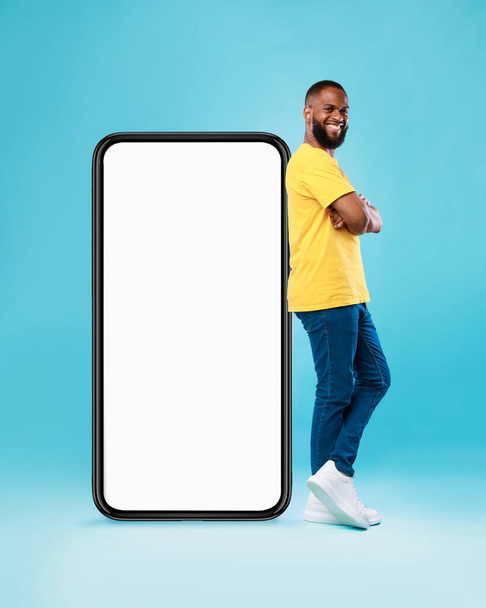 Hot offer, online sale. Funky black young guy standing near huge cellphone with mockup space over blue background - Fotografie, Obrázek