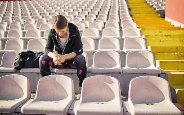 Una joven está sentada en la tribuna y descansando después de un entrenamiento en un hermoso día en el estadio. Deporte, atletismo, atletas - Foto, imagen