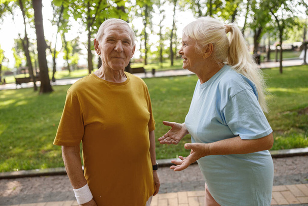 Active mature woman talks to positive man at training in green city park - Photo, Image