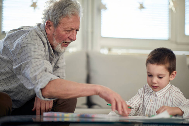 A grandpa teaching his grandson how to draw while spending a time in a relaxed atmosphere at home together. Family, home, playtime - Photo, image