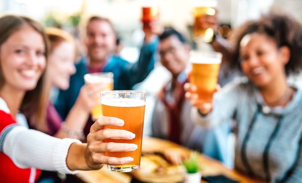 Blurred background of friends drinking and toasting beer at brewery bar restaurant patio - Happy hour promotion concept with people having fun together at brew out door pub - Focus on lower pint glass - 写真・画像
