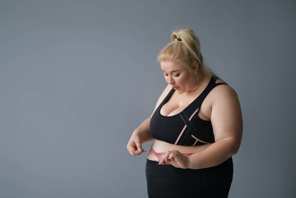 Νεαρή γυναίκα με παραμορφωμένο πρόσωπο μετράει το μέγεθος της κοιλιάς με μεζούρα. Αστεία ξανθιά παχύσαρκη γυναίκα σε ένα σπορ μαύρο κοστούμι, απομονώνονται σε ένα γκρι φόντο.Δίαιτα δίνει ένα θετικό αποτέλεσμα - Φωτογραφία, εικόνα