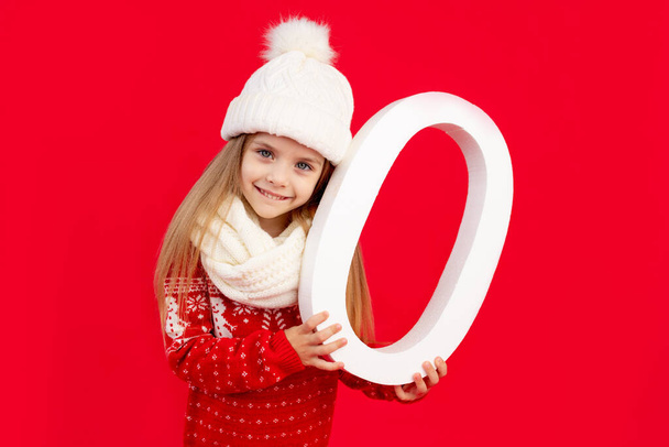 dziewczynka w czapce zimowej i swetrze z dużą liczbą zero na czerwonym, monochromatycznym tle raduje się i uśmiecha, koncepcja Nowego Roku i Bożego Narodzenia, przestrzeń na tekst - Zdjęcie, obraz