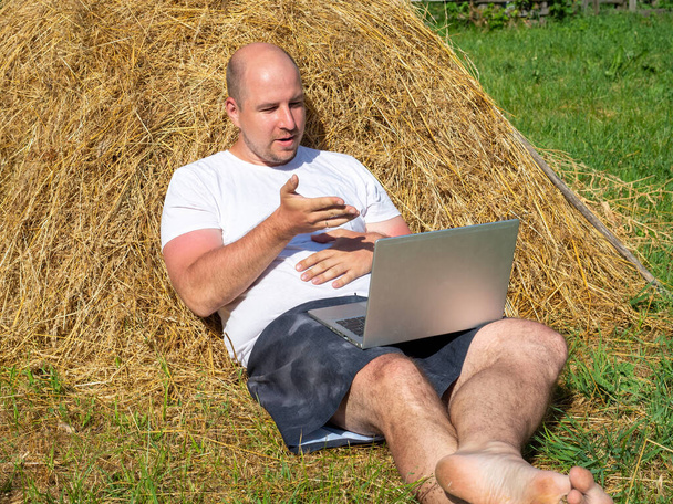 Tişört ve şort giymiş orta yaşlı bir adam elinde gri bir dizüstü bilgisayarla sarı bir samanlıkta yatıyor. Uzaktan çalışma, eğitim. Kırsal bölge. Yüzündeki öfke, duygular - Fotoğraf, Görsel