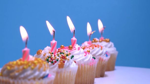 Birthday cupcakes with burning candles on blue background. Vanilla coconut muffins with white whipped cream and sugar sprinkles icing. Close-up shot with shallow depth of field. - Foto, afbeelding