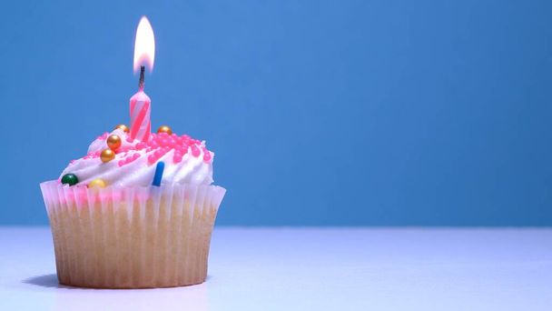 Cupcake with single birthday candle burning and sugar sprinkles icing on blue background. Delicious muffin decoration. Homemade vanilla cup cake with buttercream frosting. Shallow depth of field. - Foto, afbeelding