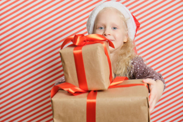 cute cheerful preschooler girl in Santa hat with gifts on red striped background, gifts with a red ribbon in the hands of girl with blond hair, boxing day, birthday gifts and christmas, holiday gifts - 写真・画像