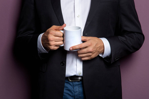 a man in a black jacket holds a white mug of tea in his hands. Free space on the mug for the inscription. concept is a sample for a corporate logo. - Photo, Image