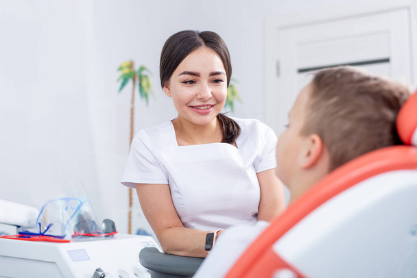 female dentist talks to a young boy and tells him how to take care of his teeth. handsome boy is smiling in dentist's office. concept is a children's medical examination. - Zdjęcie, obraz