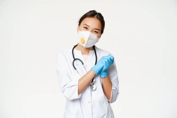 Sonriendo lindo médico asiático, enfermera en respirador médico y guantes estériles de goma, mirando complacido en la cámara, de pie en uniforme de trabajador de la salud, fondo blanco - Foto, Imagen