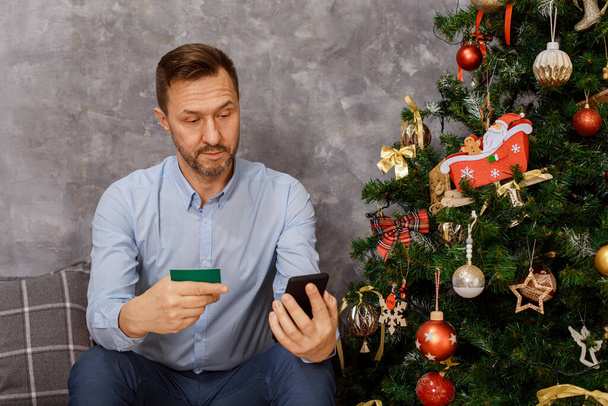 man zit in de buurt van kerstboom met mobiele telefoon, smartphone en creditcard in handen, hij is geschokt door de kosten van geschenken, emoties, verrassing, ontevredenheid.  - Foto, afbeelding