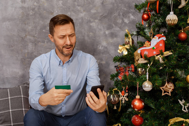 Mann sitzt am Weihnachtsbaum mit Handy, Smartphone und Kreditkarte in der Hand, er ist schockiert über die Kosten für Geschenke, Emotionen, Überraschung, Unzufriedenheit.  - Foto, Bild