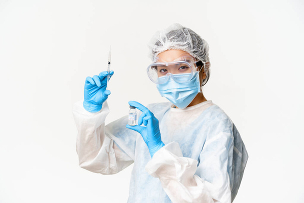 Азиатский врач или медсестра в медицинском защитном оборудовании, уверенно смотрящие в камеру, держащие шприц и вакцину от ковида-19, кампанию вакцинации и медицинскую концепцию - Фото, изображение