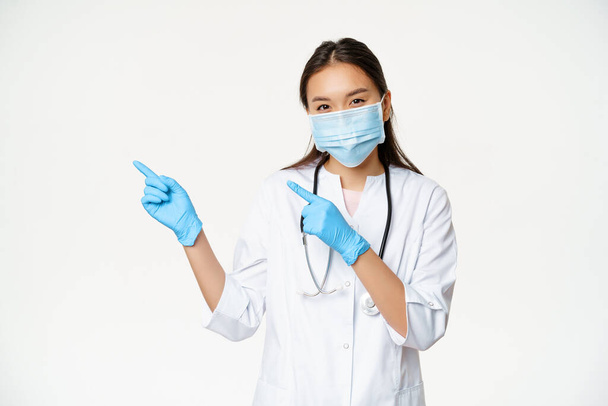 Retrato de una trabajadora médica asiática señalando con los dedos hacia la izquierda, usando mascarilla y guantes de goma, de pie en uniforme de la clínica sobre fondo blanco - Foto, Imagen