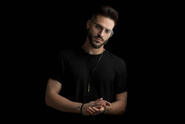 confident fashion man in black outfit wearing glasses and rubbing palms while posing on black background in studio - Photo, Image