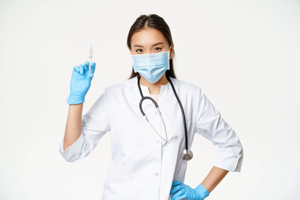 Азійська медсестра в медичній масці зі шприцом з вакциною, концепцію охорони здоров'я і вакцинації, стоячи в клінічній формі, білий фон - Фото, зображення