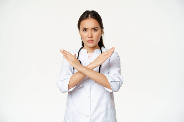 Серйозна азіатська жінка-лікар забороняє поведінку пацієнта, показуючи зупинку, жест хрестоподібних рук, замерзлий незадоволений, не схвалює поганий смоктання, стоїть на білому тлі
 - Фото, зображення