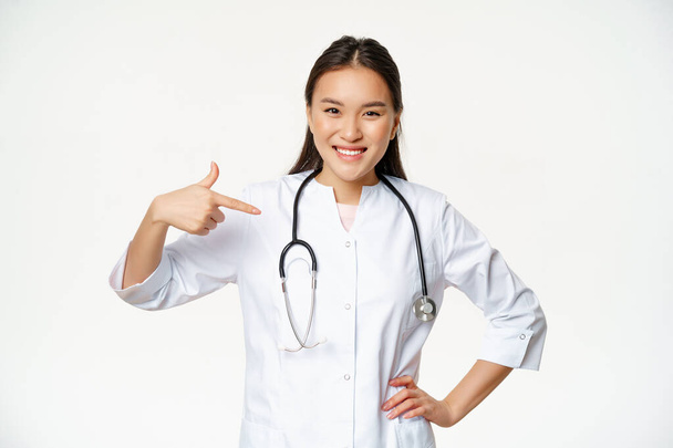 Smiling asian female doctor, real professional medical worker, pointing fingers at herself, wearing medical robe and stethoscope, white background - Photo, Image