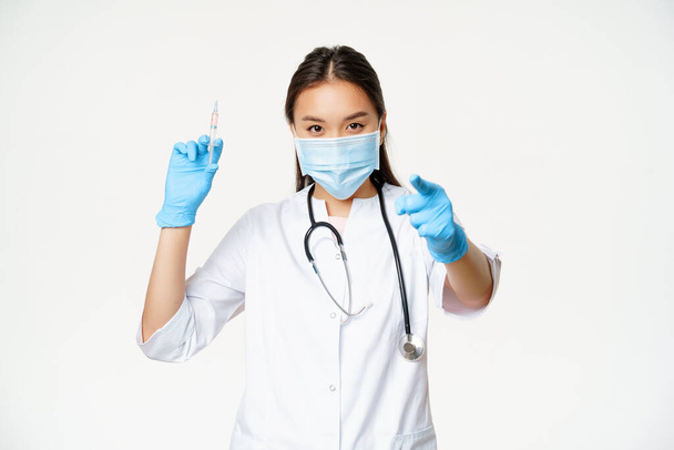 Szczepienie koronawirusem. Lekarz azjatycki, lekarz trzymający strzykawkę ze szczepionką i wskazujący na pacjenta przed kamerą, kampania szczepień, białe tło - Zdjęcie, obraz