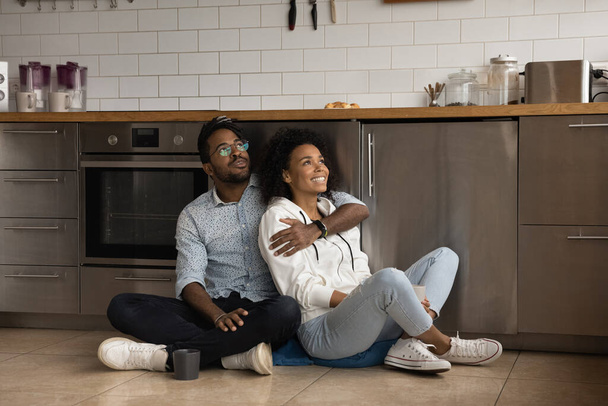 Happy young mixed race couple sitting on floor in kitchen. - Φωτογραφία, εικόνα