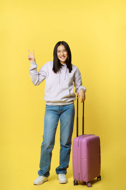 De cuerpo entero tiro feliz chica asiática, turista con maleta va de vacaciones, sonriendo y mostrando signo de paz, fondo amarillo - Foto, Imagen