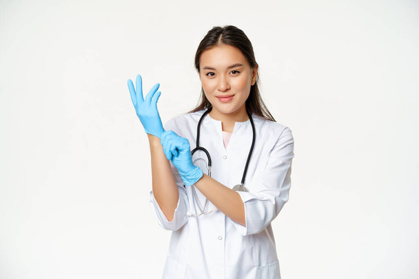 Gülümseyen, kendine güvenen Asyalı kadın doktor tıbbi eldiven takıyor, hasta muayenesine hazırlanıyor, beyaz arka planda üniformayla dikiliyor. - Fotoğraf, Görsel
