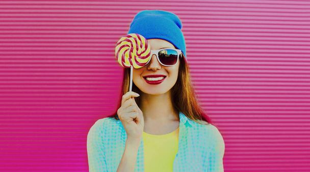 Portrait of smiling young woman with sweet colorful lollipop on pink background - 写真・画像