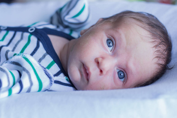 A small baby with blue eyes in a striped suit lies on a white pillow and looks slightly to the side of the frame. - Foto, immagini