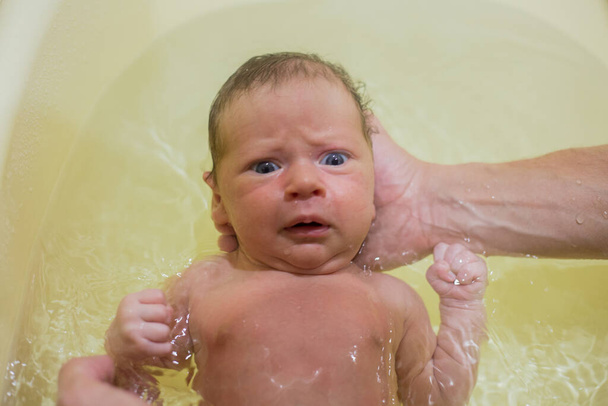 A newborn baby is bathing in a bath for the first time, his hand is holding his head, the baby is scared. - Foto, imagen