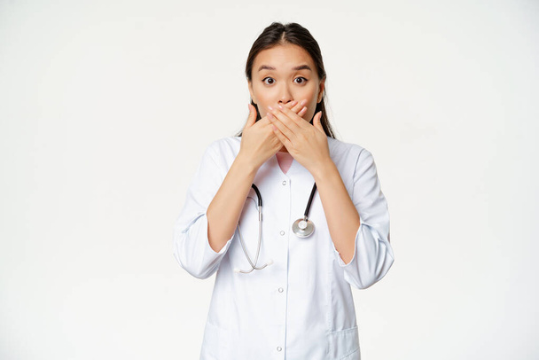 ショックを受けた女性のアジアの医師の画像は、手で彼女の唇をカバー,口を閉じて、心配そうな,タブージェスチャー,白い背景の上に立って - 写真・画像