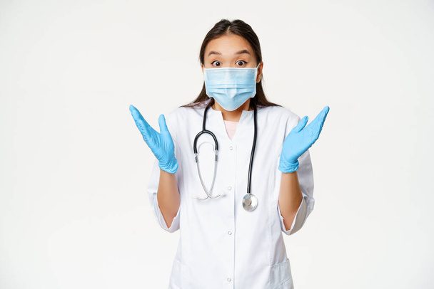 Retrato de la mujer asiática feliz médico aplauden las manos, mirando entusiasta en noticias sorprendentes, usando máscara médica y guantes de goma, fondo blanco - Foto, Imagen